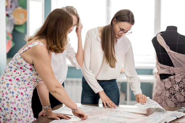 Drie knap uitziende mooie vrouwen wijzen naar de kleermakersworkshop van knipmode