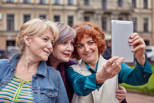 Drie lachende oudere blanke vrouwen maken selfie in het centrum van de Europese stad met tabletcomputer.