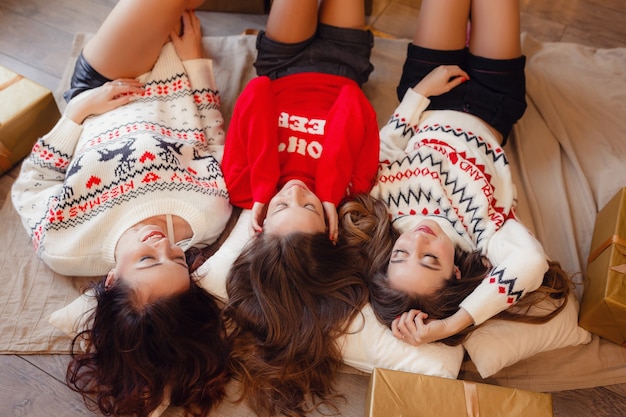 Drie mooie meisjes liggen in de buurt van de kerstboom te wachten op geschenken. Jonge vrouw beste vrienden die Kerstmis in huis vieren. Prachtige gouden kerstversiering op een hoge kerstboom
