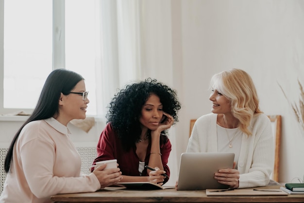 Drie mooie volwassen vrouwen gebruiken een digitale tablet terwijl ze samen aan het bureau zitten.