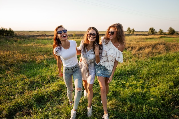 Drie prachtige jonge vrouwen in zonnebril gekleed in de mooie kleren staan in het veld en glimlachen op een zonnige dag. .