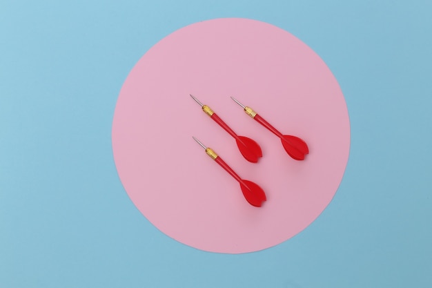 Drie rode plastic darts met metalen punt op blauwe pastelachtergrond met roze cirkel.