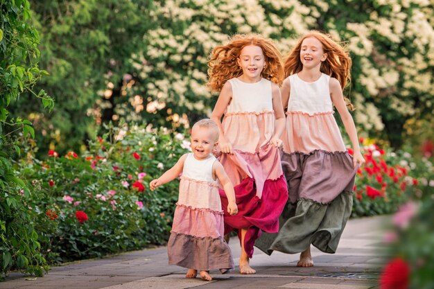 Drie roodharige zussen in lange linnen jurken lopen op zomeravond vrolijk in de tuin met rozen