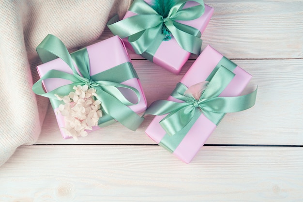 Drie roze geschenkdozen met linten en een trui op een witte houten achtergrond.