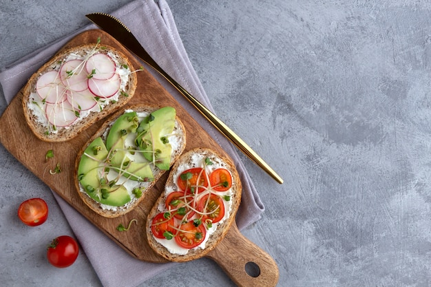 Drie sandwiches op een snijplank met graan brood, groenten en microgreens op een grijze achtergrond