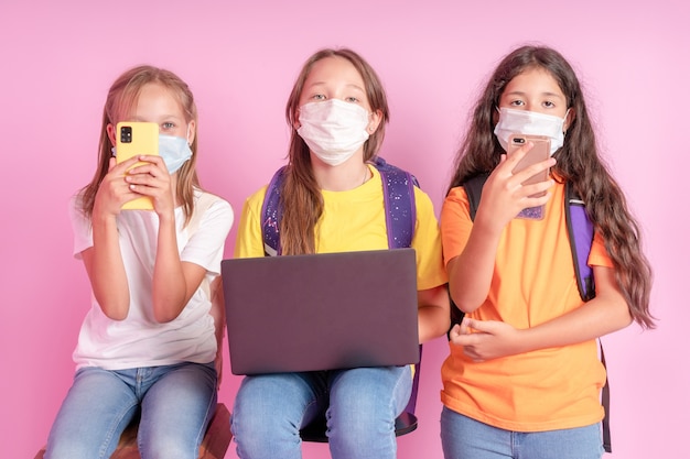 Drie schoolmeisjes in medische maskers met telefoons en een laptop in hun handen kijken naar de camera op een roze achtergrond.