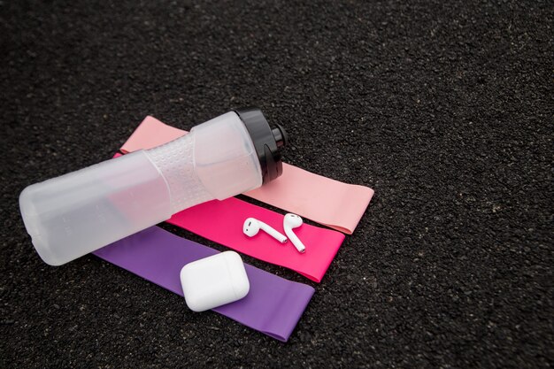 Drie soorten fitnessgombanden voor sport, draadloze hoofdtelefoons en een fles water op een zwarte muur
