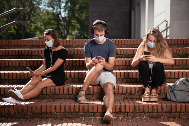 Drie studenten die op de trap zitten en hun smartphones gebruiken met een gezichtsmasker op