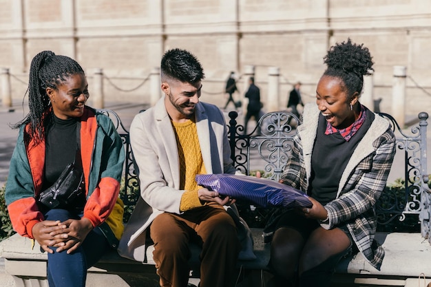 Drie vrienden van verschillende etniciteiten wisselen geschenken uit terwijl ze buiten op een bankje zitten