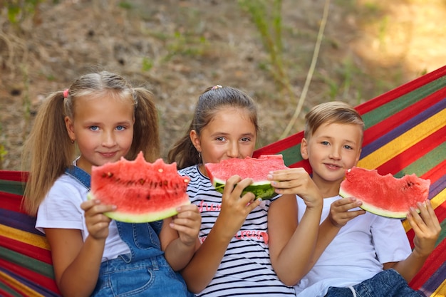 Drie vrolijke kinderen eten watermeloen en grappen, buiten, zittend op een kleurrijke hangmat. zomerplezier en ontspanning