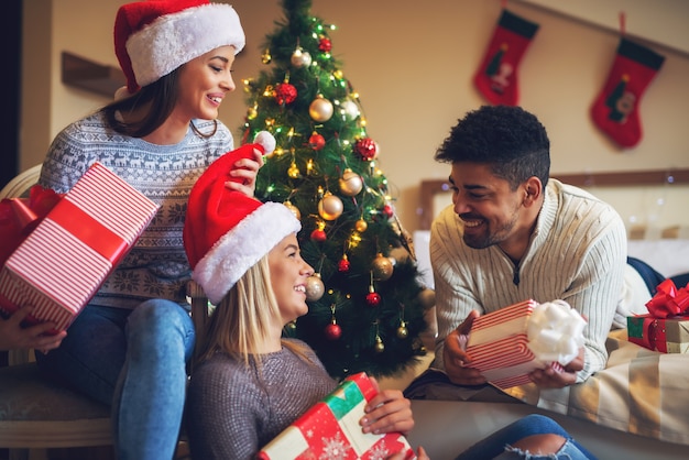 Drie vrolijke stijlvolle vrienden met kerstmutsen in truien met cadeautjes en grappen praten met elkaar in de slaapkamer terwijl ze thuis genieten van de kerstvakantie.