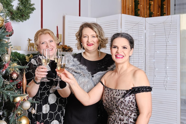 Drie vrolijke vrouwen van middelbare leeftijd die Kerstmis vieren terwijl ze in de buurt van de kerstboom staan