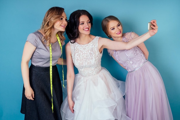 Drie vrouwen in de kleedkamer. naaister vrouw en twee bruid in trouwjurk ido selfie n de studio op een blauwe achtergrond. kleermaker en bruidsmeisjes