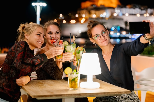 Drie vrouwen maken een selfie terwijl ze op een terras zitten en 's nachts cocktails drinken