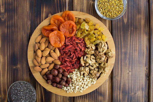 Dried apricots, raisins, goji berries,different nuts, chia seeds and  bee pollen on the round cutting board on the brown  wooden background.Top view.