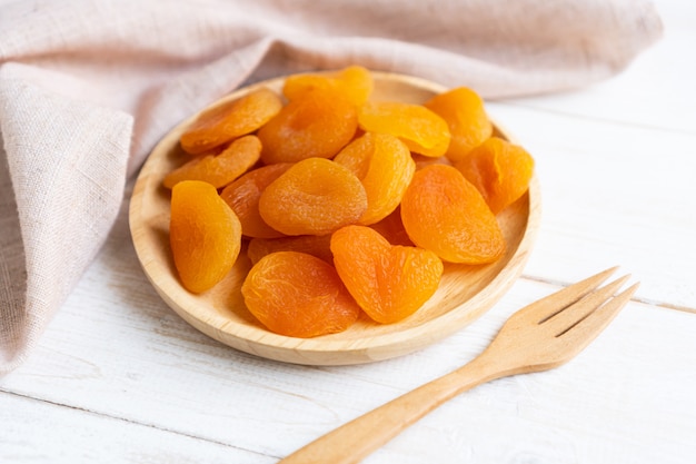 Dried apricots in wooden plate on white wooden table with table cloth and fork, close up.