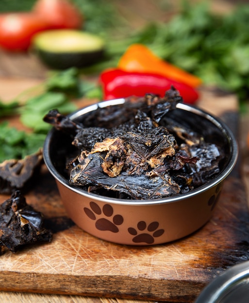 Photo dried beef liver in the dog vessel among the greenery on the wooden board and some vegetables on the background. chewing treats for domestic dogs.