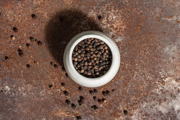 Dried black peppercorns in a ceramic bowl. Rusty metal background, close up. Trendy hard light, dark shadow, top view