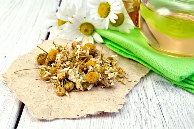 Dried chamomile flowers on paper, tea in glass teapot on a napkin, fresh daisy flowers on a background of pale wooden plank