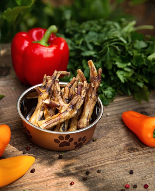 Cosce di pollo essiccate nella nave per cani tra il verde sulla tavola di legno e alcune verdure sullo sfondo. dolcetti da masticare per cani domestici.