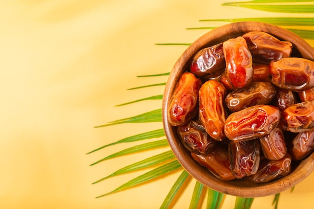 Dried dates in wooden bowl with palm leaves on yellow background. Top view. Space for text.