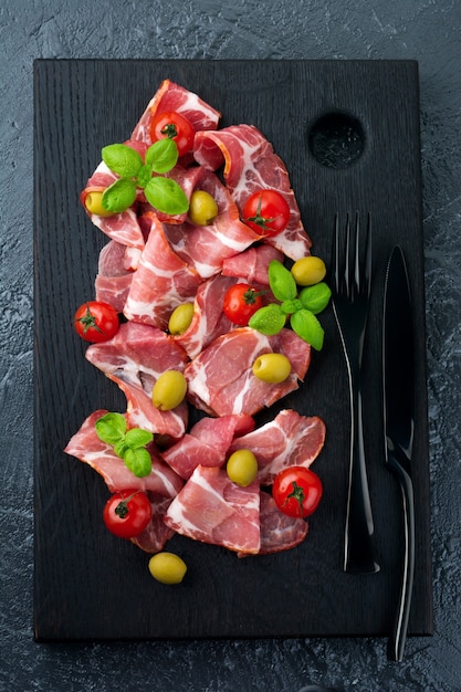 Dried meat, jamon with cherry tomatoes, basil and olives on a black 