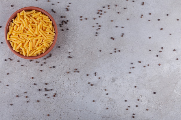 Dried penne Italian pasta in a round bowl with pepper corns on a stone table .