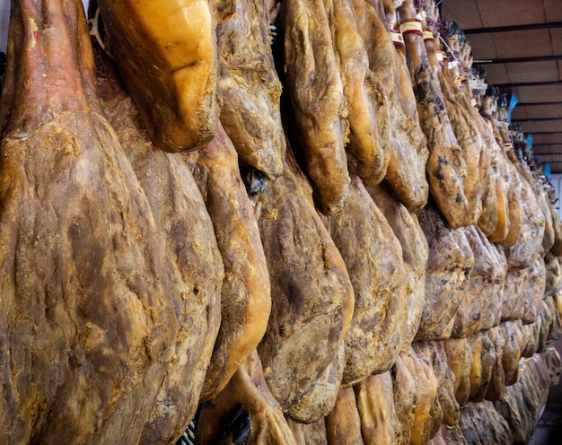 На мясном рынке висят сушеные свиные бедра. Испанское национальное блюдо из ветчины или хамона в продуктовом магазине. Покупки иберийской свинины в супермаркете Испании. Сушеный и вяленый окорок на развешивании. Рынок продают сырые мясные продукты