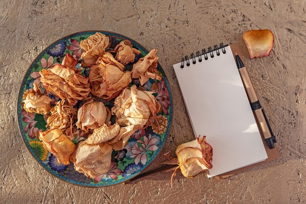 Dried Rose flowers On Old Vintage plate in sunlight. Dry roses heads on lit by the sun and a notepad with pan for scrapbook. Copy space.