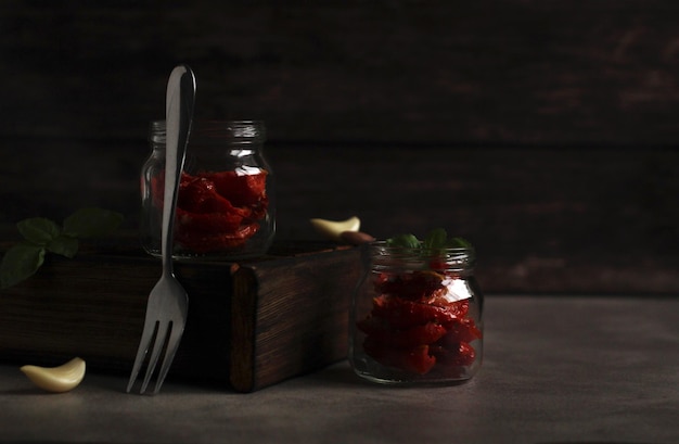 Dried tomatoes in a glass jar with garlic and basil with a fork on a dark background