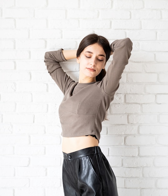 Driekwart lengte portret van mooie lachende brunette vrouw met lang haar bruin shirt en zwarte lederen broek, dragen op witte bakstenen muur achtergrond