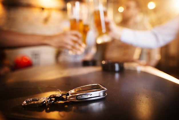 Don't drink and drive! Car key on a wooden pub table in front of blurred friend clinking with a beer.