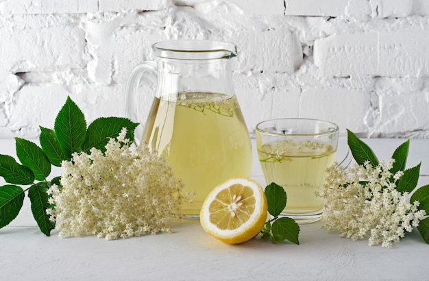 Drink from elderberry flowers in carafe with slice of lemon against  white brick wall.