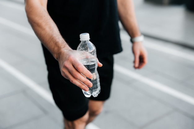 Выпей немного воды! Рука человека протягивает пластиковую бутылку с водой. Серый асфальт на фоне. Человек в черной спортивной беге трусцой. Время гидратации! Тренировка на свежем воздухе.