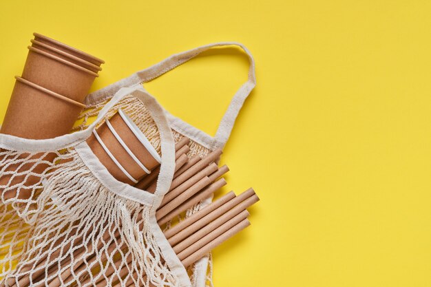 Drinking brown tubes straws made of paper and cornstarch, mesh market bag and empty paper coffee cups on a trendy grey and yellow background. Zero waste and plastic free concept. Top view.