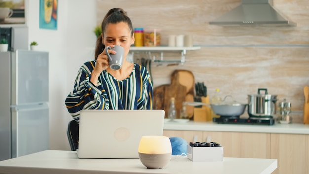 Drinking coffe and working with aromather apy essential oils diffuser in the kitchen. Aroma health essence, welness aromatherapy home spa fragrance tranquil theraphy, therapeutic steam, mental health