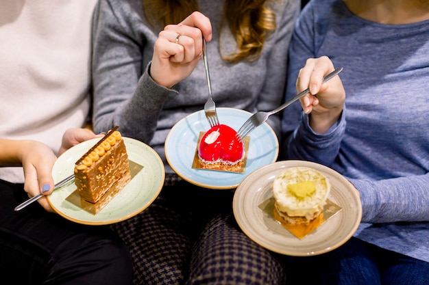 Drinking coffee and eating desserts together. Top view of hands of three beautiful women holding plates with delicious cakes desserts in cafe. Meeting of best friends. Coffee with cakes