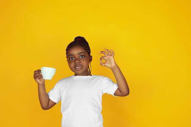 Drinking coffee, tea, shows nice gesture. Little african-american girl's portrait on yellow studio background. Cheerful kid. Concept of human emotions, expression, sales, ad. Copyspace. Looks cute.