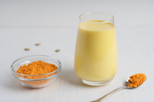 Drinking glass full of golden milk served with spoon of turmeric on white wooden table