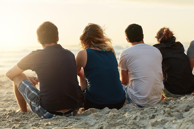 Вместе пьют, наслаждаясь видом. Снимок заднего вида группы неопознанных друзей, которые вместе любуются закатом, сидя на пляже.