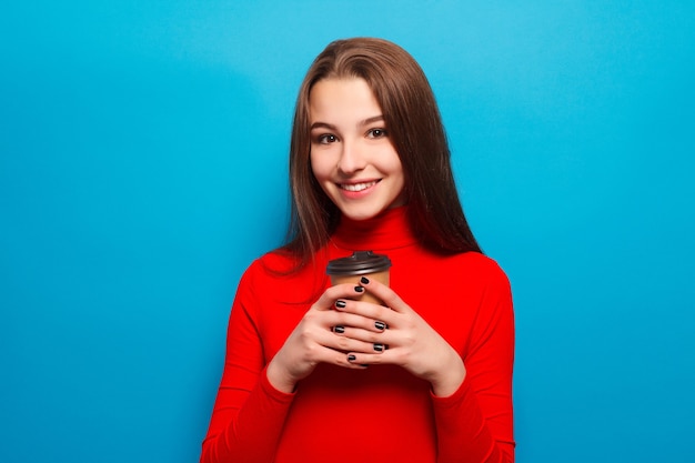 Drinks, emotions, people, beauty, lifestyle concept - emotional happy beautiful woman in red blouse on blue studio background drinking coffee in paper cup smiling, positive, enjoying, morning food