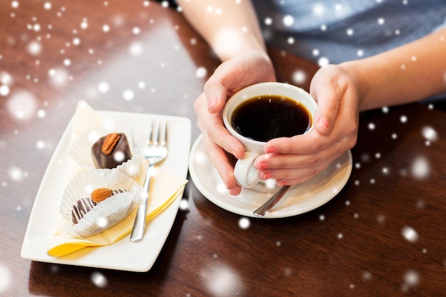 drinks, people and lifestyle concept - close up of woman hands holding cup with hot black coffee and dessert over snow