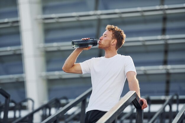 Drinks water and takes a break Young man in sportive clothes have workout outdoors at daytime