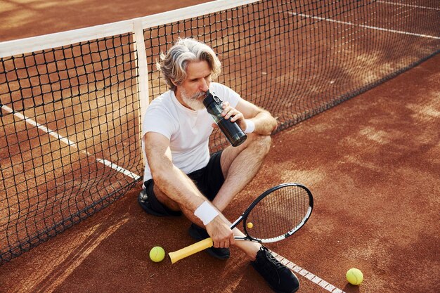 Drinkwater en zittend op de grond Senior moderne stijlvolle man met racket buitenshuis op tennisbaan overdag