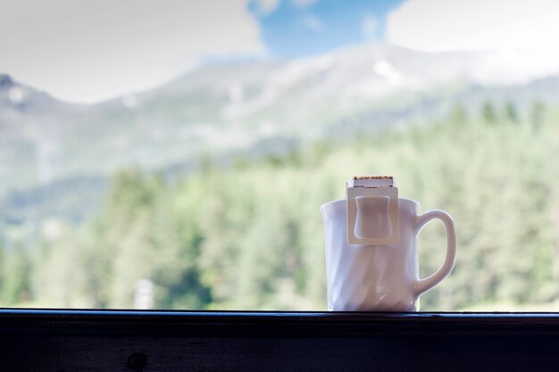 コーヒーを淹れる山の風景のトレンドを背景に、マグカップにコーヒーバッグを滴下します。