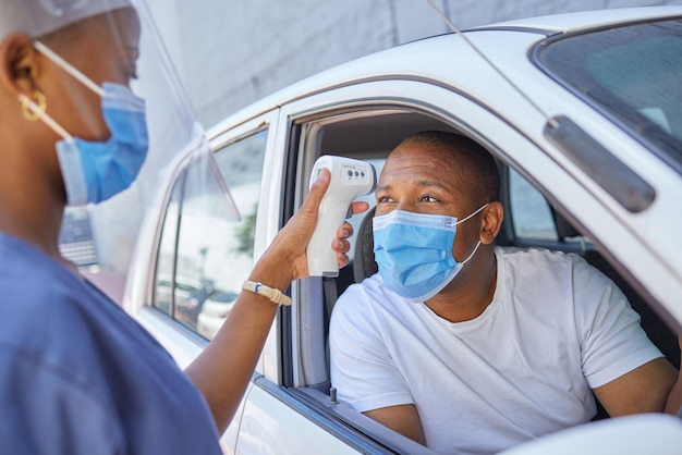 Drive thru for covid screening or testing service for people driving or traveling Black man wearing a mask in a car getting his temperature test using an infrared thermometer as coronavirus protocol