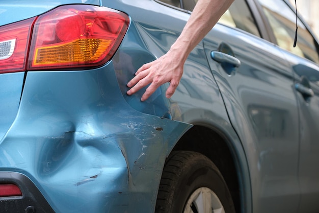 Driver hand examining dented car with damaged fender parked on\
city street side road safety and vehicle insurance concept