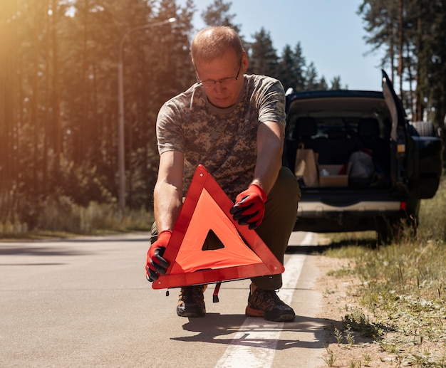 Фото Водитель мужчина ставит знак осторожности треугольник на дороге возле разбитого автомобиля летом