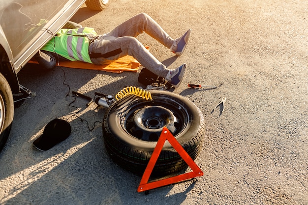 A driver or worker repairs a broken car on the side of the road. View from above. Man is under the car