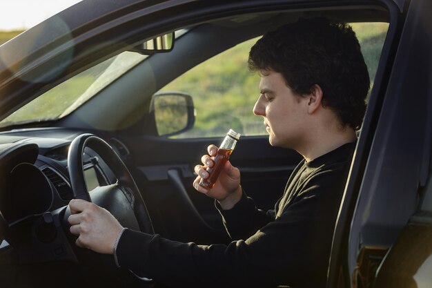 車を運転している若い男の運転手は、ガラス瓶からアルコール飲料を飲みます